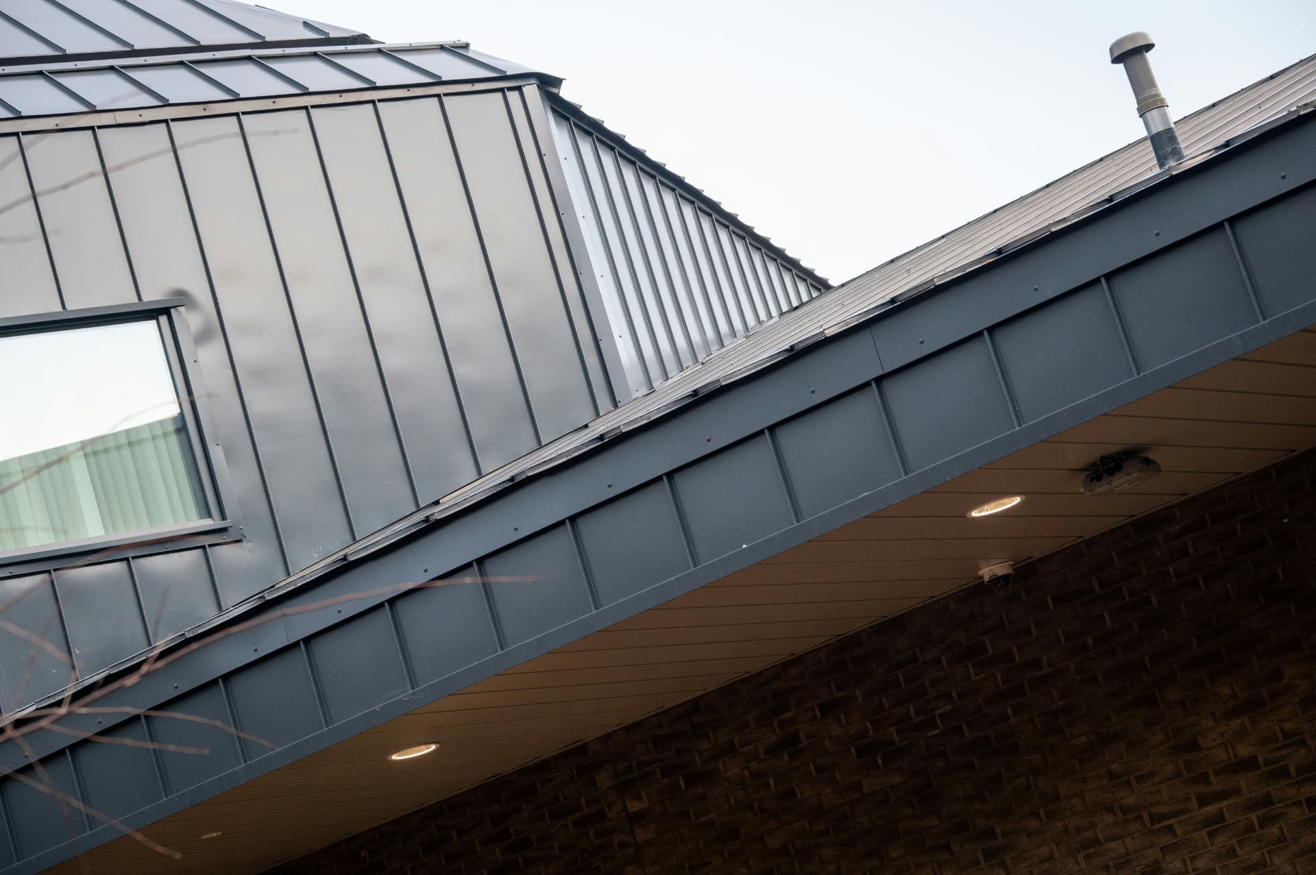 Angled dark standing-seam metal roof planes with a vent, window and brick wall